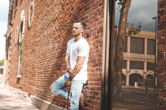 A person in a white t-shirt and ripped jeans is leaning against a brick wall. The scene is urban, with the reflection of a tree and buildings visible in a large window. The image highlights casual fashion and has a relaxed atmosphere.