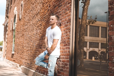 A person in a white t-shirt and ripped jeans is leaning against a brick wall. The scene is urban, with the reflection of a tree and buildings visible in a large window. The image highlights casual fashion and has a relaxed atmosphere.