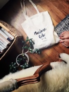 A white tote bag with 'Marlene Coto' written on it is placed on a wooden floor beside a knitted rug, a houseplant in a pot, and a wicker basket filled with books. A person's bare feet with painted toenails are partially visible near the rug.