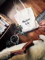 A white tote bag with 'Marlene Coto' written on it is placed on a wooden floor beside a knitted rug, a houseplant in a pot, and a wicker basket filled with books. A person's bare feet with painted toenails are partially visible near the rug.