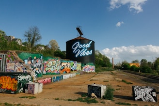 A large cylindrical structure with neon text reading 'Good Vibes' is prominently displayed, surrounded by colorful graffiti on nearby walls and surfaces. The scene includes train tracks running alongside the graffiti-covered area, with trees and a bright blue sky providing a natural backdrop.