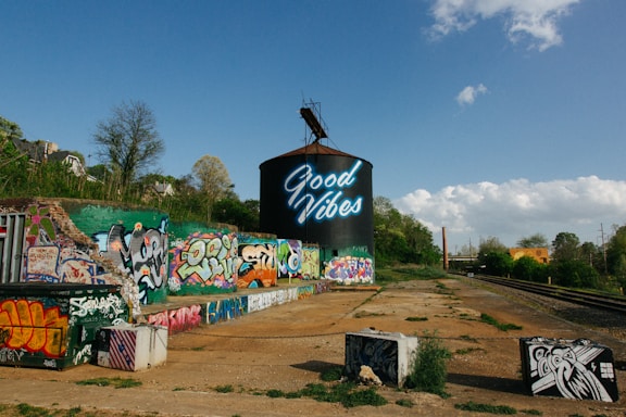 A large cylindrical structure with neon text reading 'Good Vibes' is prominently displayed, surrounded by colorful graffiti on nearby walls and surfaces. The scene includes train tracks running alongside the graffiti-covered area, with trees and a bright blue sky providing a natural backdrop.