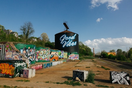 A large cylindrical structure with neon text reading 'Good Vibes' is prominently displayed, surrounded by colorful graffiti on nearby walls and surfaces. The scene includes train tracks running alongside the graffiti-covered area, with trees and a bright blue sky providing a natural backdrop.