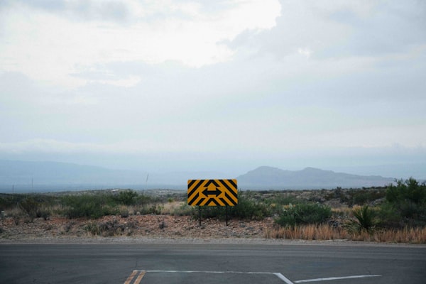 a yellow-and-block road sign with two arrows pointing to the side, against a mountainous backdrop