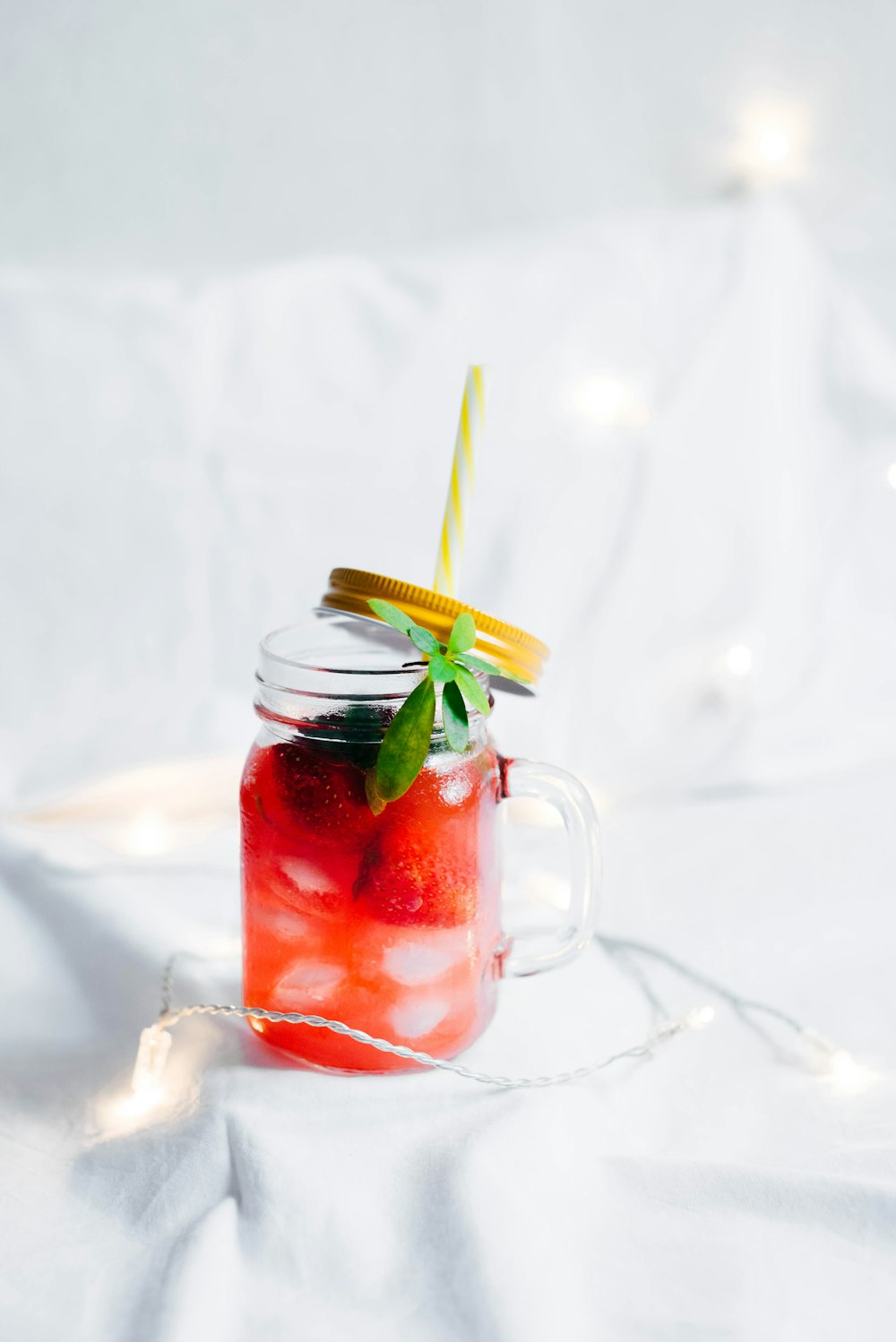 Tasse Mason en verre transparent avec boisson à la fraise sur textile blanc entouré d’une guirlande lumineuse