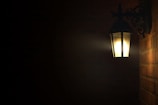 A dimly lit outdoor wall-mounted lantern illuminates a surrounding brick wall. The light casts a warm glow in an otherwise dark space, with intricate metalwork supporting the fixture.