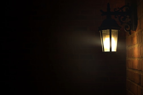 A dimly lit outdoor wall-mounted lantern illuminates a surrounding brick wall. The light casts a warm glow in an otherwise dark space, with intricate metalwork supporting the fixture.