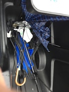 A collection of broken umbrellas is tangled together in the corner of a dark, slick surface, likely the inside of a trunk. The umbrellas have exposed frames with bent or snapped ribs, and fabric in blue and plaid patterns. A noticeable orange handle is present.