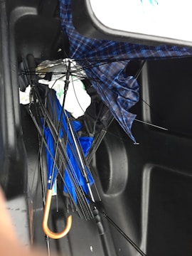 A collection of broken umbrellas is tangled together in the corner of a dark, slick surface, likely the inside of a trunk. The umbrellas have exposed frames with bent or snapped ribs, and fabric in blue and plaid patterns. A noticeable orange handle is present.