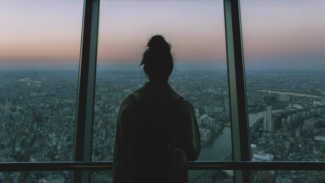 Silhouette of a person standing at a floor-to-ceiling window, looking out over a city at dusk