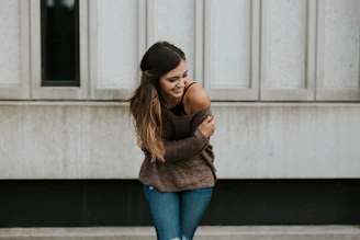 Confident woman smiling softly, wearing a warm nude-toned hoodie, standing against a minimal cream wall.