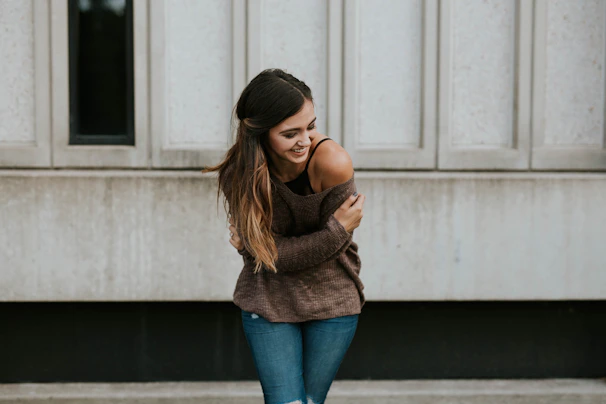 Confident woman smiling softly, wearing a warm nude-toned hoodie, standing against a minimal cream wall.