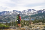 A hiker stands on a rocky outcrop overlooking a vast mountainous landscape. Snow-capped peaks rise in the distance, surrounded by dense green forests. Below, a clear lake reflects the sky. The hiker carries a green backpack and wears outdoor gear suited for hiking.
