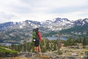 A hiker stands on a rocky outcrop overlooking a vast mountainous landscape. Snow-capped peaks rise in the distance, surrounded by dense green forests. Below, a clear lake reflects the sky. The hiker carries a green backpack and wears outdoor gear suited for hiking.