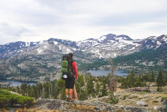 A hiker stands on a rocky outcrop overlooking a vast mountainous landscape. Snow-capped peaks rise in the distance, surrounded by dense green forests. Below, a clear lake reflects the sky. The hiker carries a green backpack and wears outdoor gear suited for hiking.