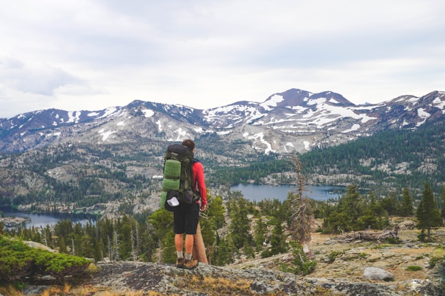 A hiker stands on a rocky outcrop overlooking a vast mountainous landscape. Snow-capped peaks rise in the distance, surrounded by dense green forests. Below, a clear lake reflects the sky. The hiker carries a green backpack and wears outdoor gear suited for hiking.