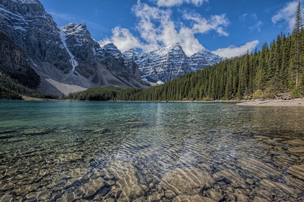Crystal-clear mountain lake reflecting deep green forest and soft blue sky.