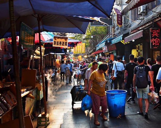 A bustling street market lined with vibrant stalls and awnings in various colors. People walk through the narrow street, while others stand examining goods. Signs with Chinese characters hang in the background, and a few bicycles can be seen. Steam rises from one of the stalls, creating a lively and busy atmosphere.