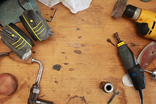 A wooden workbench scattered with various tools including a pair of ear protectors, a cordless drill, a rotary tool with attachments, a small wrench, and other metal and wood components. The tools are arranged in a disorganized manner, suggesting a busy workspace.