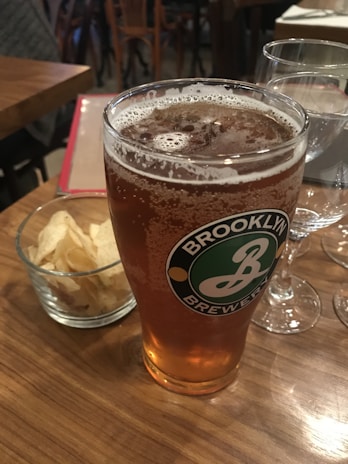 A glass of amber beer with a Brooklyn Brewery logo is placed on a wooden table, accompanied by a small bowl of potato chips and a wine glass. The setting appears to be in a casual dining environment with wooden furniture visible in the background.