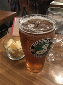 A glass of amber beer with a Brooklyn Brewery logo is placed on a wooden table, accompanied by a small bowl of potato chips and a wine glass. The setting appears to be in a casual dining environment with wooden furniture visible in the background.