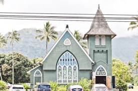 A quaint church with a pointed tower and stained glass windows stands amidst a lush tropical landscape. Palm trees surround it, and a mountain range is visible in the background. Several cars are parked in front of the church beneath a set of power lines.