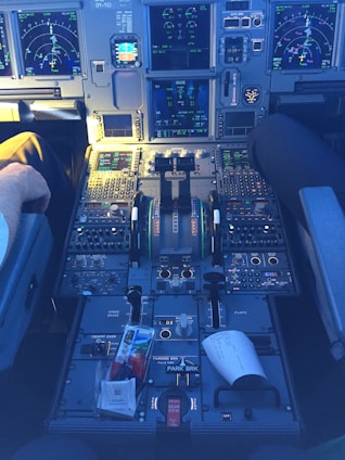 The cockpit of an aircraft, featuring a highly detailed control panel with numerous switches, buttons, and multiple digital screens displaying various flight data. Two pilots' seats are visible, with one occupied by a person. A paper and some items are seen on the central console.