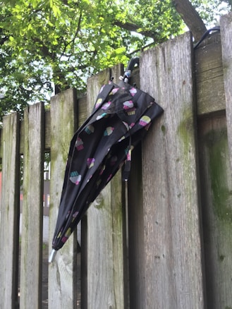 A closed umbrella with a colorful cupcake pattern is hanging over a weathered wooden fence. The fence is surrounded by lush green trees and foliage, creating a natural and serene setting.