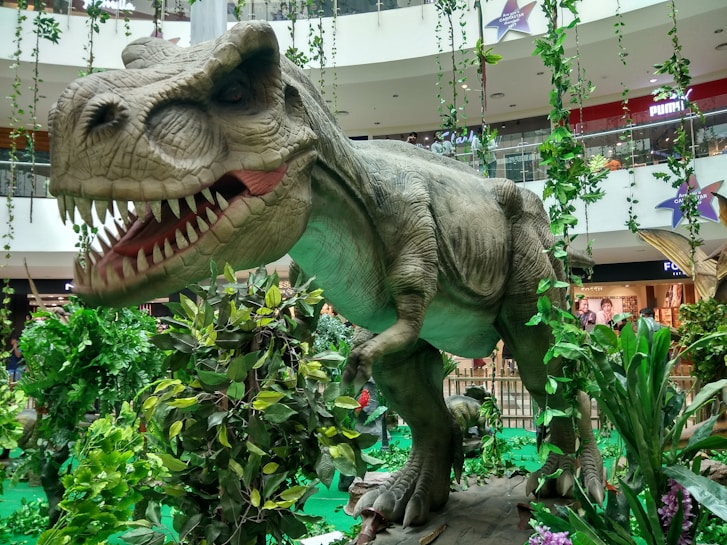 A large, realistic dinosaur model is displayed amidst lush green foliage inside a modern shopping mall. The dinosaur, resembling a T-Rex, has its mouth open showing its sharp teeth, creating an imposing presence. Surrounding it are various plants and vines, giving the scene a prehistoric jungle appearance. Shoppers can be seen in the background behind the dinosaur display area.