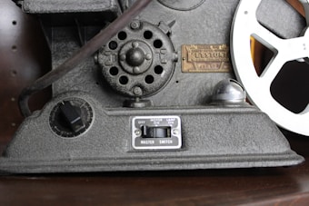 A vintage film projector featuring a metallic body with a motor speed control dial, a master switch for operation, and an intricately designed film reel. The machine displays a brass plate with the model number and branding.
