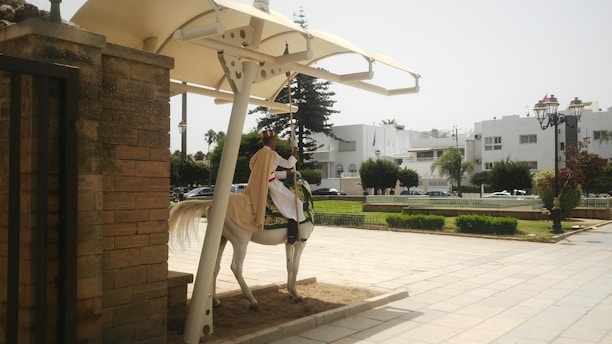 A person wearing traditional attire rides a white horse under a canopy. The setting includes a paved area surrounded by greenery and white buildings in the background. A brick wall stands to the left, and a decorative street lamp is visible.