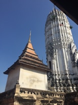 A historical temple structure with intricate architectural details is set against a clear blue sky. The temple features a tall spire adorned with traditional design elements and ornate carvings. The foreground includes a secondary building with a tiered roof, accented with decorative ridges and an aged appearance.