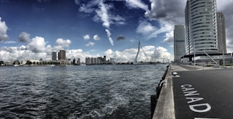 A wide river flows through an urban landscape with several modern buildings and a distinctive cable-stayed bridge in the distance. The sky is partly cloudy, with patches of blue and white clouds scattered across. The foreground features a pathway marked with the word 'CANADA' along the water's edge.