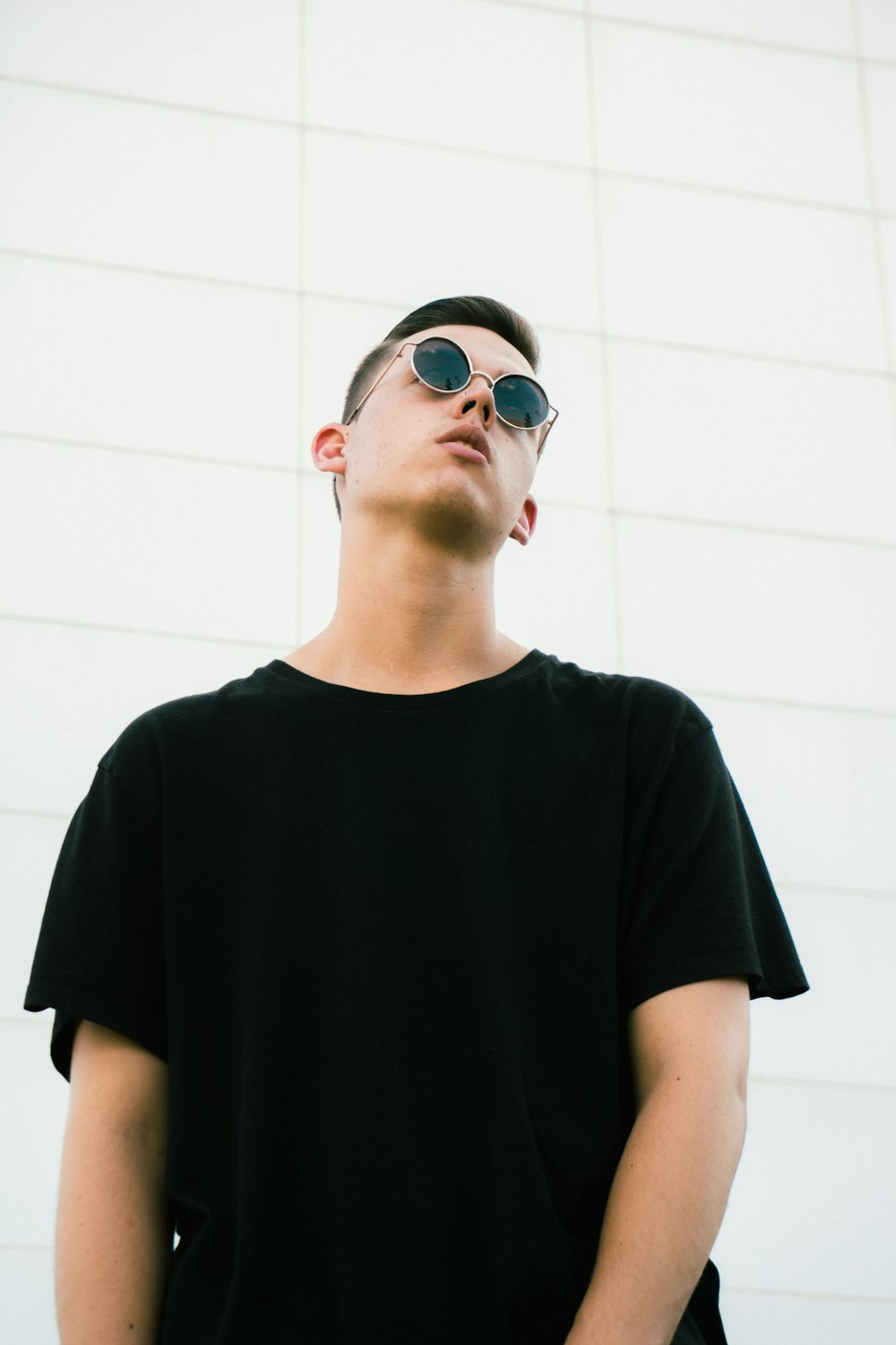 Homme portant un t-shirt noir à col rond et des lunettes de soleil debout près d’un mur blanc