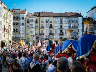 A lively street scene featuring large, intricately designed parade figures resembling medieval kings and other characters. The gathering crowd is vibrant, with spectators and participants wearing colorful clothing, indicating a festive atmosphere. Historical buildings with numerous windows serve as a backdrop.