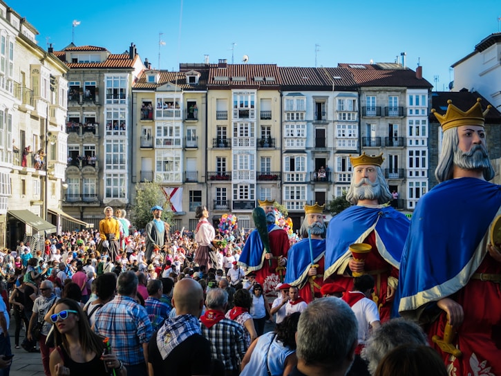 A lively street scene featuring large, intricately designed parade figures resembling medieval kings and other characters. The gathering crowd is vibrant, with spectators and participants wearing colorful clothing, indicating a festive atmosphere. Historical buildings with numerous windows serve as a backdrop.