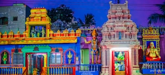 A colorful and intricately detailed temple facade featuring ornate sculptures, vibrant murals, and traditional Indian architectural elements. Statues of deities and figures adorn the entrance, with vivid blues, reds, oranges, and yellows enhancing the structure's elaborate design.