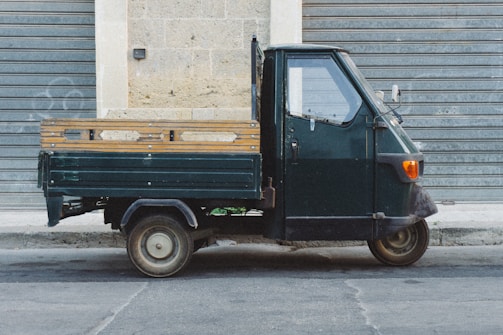 A small three-wheeled vehicle, likely a Piaggio Ape, is parked on a street. The vehicle has a dark green color and features a compact cabin with a cargo area at the back. It is positioned sideways against a metal rolling door and a stone building wall.