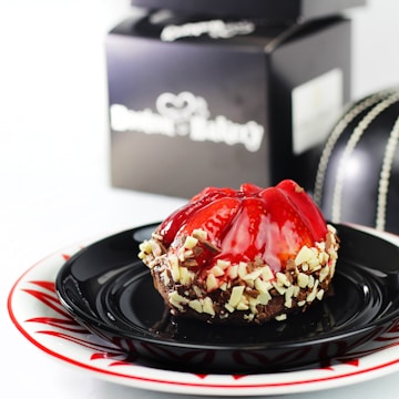 A decadent dessert is placed on a stylish black and white plate, topped with glossy red fruit glaze. The dessert is garnished with white and dark chocolate shavings around the edges. In the background, there is a blurred black box, likely packaging from a bakery.