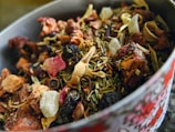 A close-up view of a bowl filled with a variety of dried herbs, fruits, and flowers. The mixture includes visible ingredients like rose petals, dried berries, and pieces of dehydrated fruit and leaves, creating a rich textural composition.