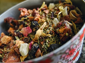 A close-up view of a bowl filled with a variety of dried herbs, fruits, and flowers. The mixture includes visible ingredients like rose petals, dried berries, and pieces of dehydrated fruit and leaves, creating a rich textural composition.
