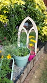 A small garden area features a mixture of vibrant plants and flowers, including yellow and purple blooms. There is a decorative, arched mirror placed among the foliage, creating the illusion of more space. The scene includes several pots in different colors, contributing to the lively and colorful appearance of the garden. A wooden border outlines the plant bed, and a patch of grass is visible nearby.