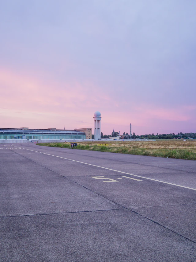 The former Tempelhof Airport runway stretches toward the airport's control tower and hangar building under a pink sunset sky.
