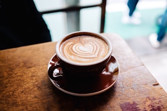 A warm latte with intricate latte art resting on a wooden table near a sunlit window in a quaint café.