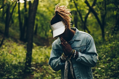 A person stands with clasped hands in a praying gesture, wearing a denim jacket and a visor cap in a lush, green forest setting. The light filters through the trees, creating a serene atmosphere.