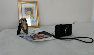 On a light-colored surface, there is a framed flower painting with a golden border in the background. A heart-shaped photo frame, a printed photograph of a child, and a black digital camera with a strap are placed in front of it.