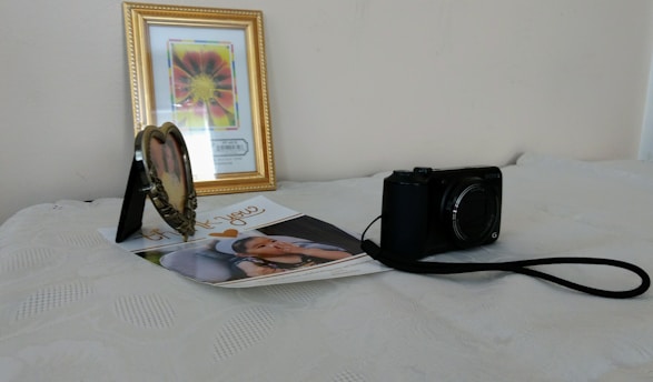 On a light-colored surface, there is a framed flower painting with a golden border in the background. A heart-shaped photo frame, a printed photograph of a child, and a black digital camera with a strap are placed in front of it.