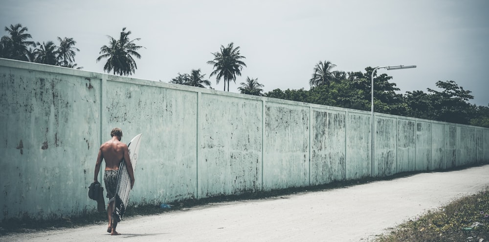 man walking on road beside wall while holding soarboard