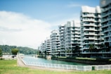 Modern apartment buildings line a waterway with several floors of balconies, surrounded by greenery and palm trees. The sky is clear with few clouds, and there is a blurred effect giving a dreamy aesthetic.
