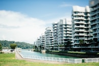 Modern apartment buildings line a waterway with several floors of balconies, surrounded by greenery and palm trees. The sky is clear with few clouds, and there is a blurred effect giving a dreamy aesthetic.